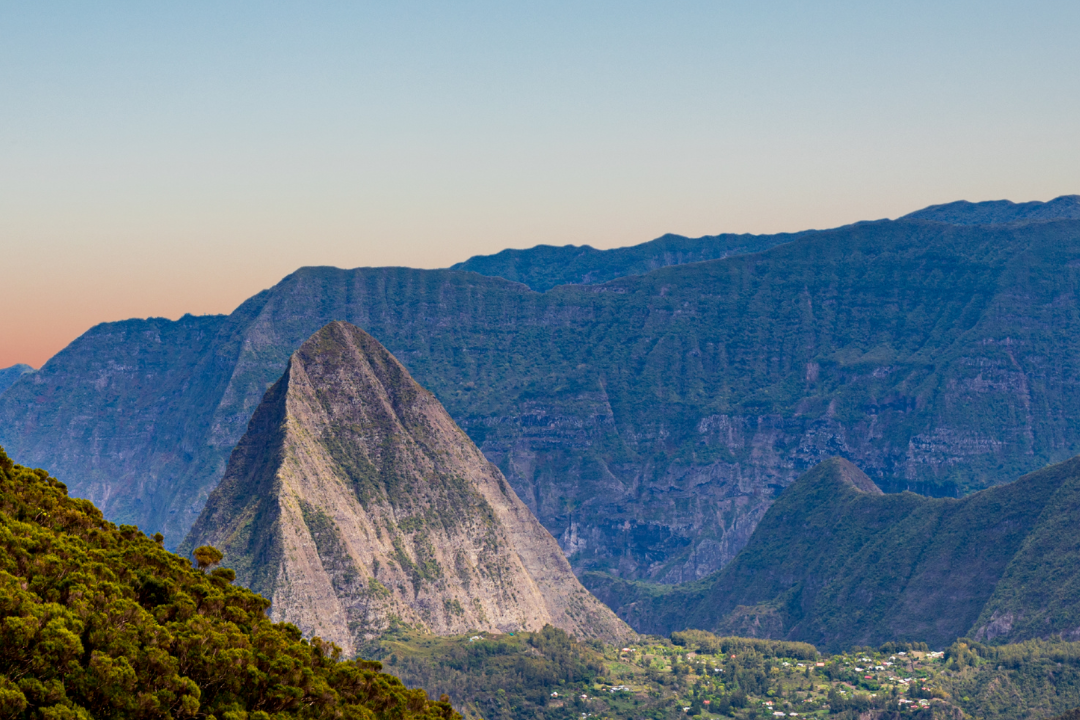 Vue panoramique sur un piton montagneux escarpé entouré de vallées verdoyantes et de falaises, avec un village niché au pied de la montagne, sous un ciel clair au coucher du soleil.